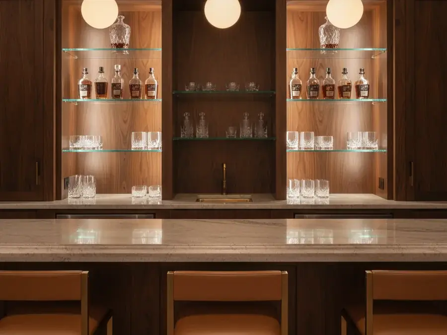 Home bar with long quartzite countertop, brass barstools, and backlit walnut shelves